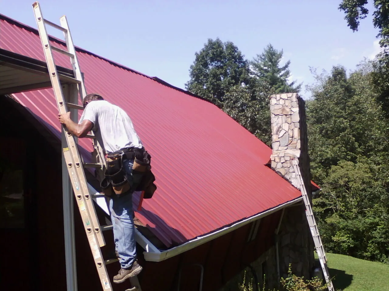 Roofer on a ladder beside a red metal roof and stone chimney — Soffit & Fascia Repair by Roofing Brothers in Dry Run